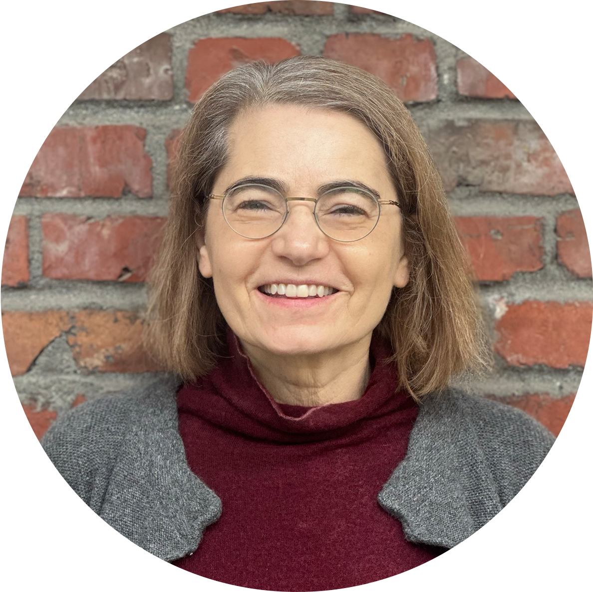 A woman with shoulder-length brown hair, wearing glasses, a gray coat with shoulder pads, and a maroon turtleneck, smiling in front of a brick wall.