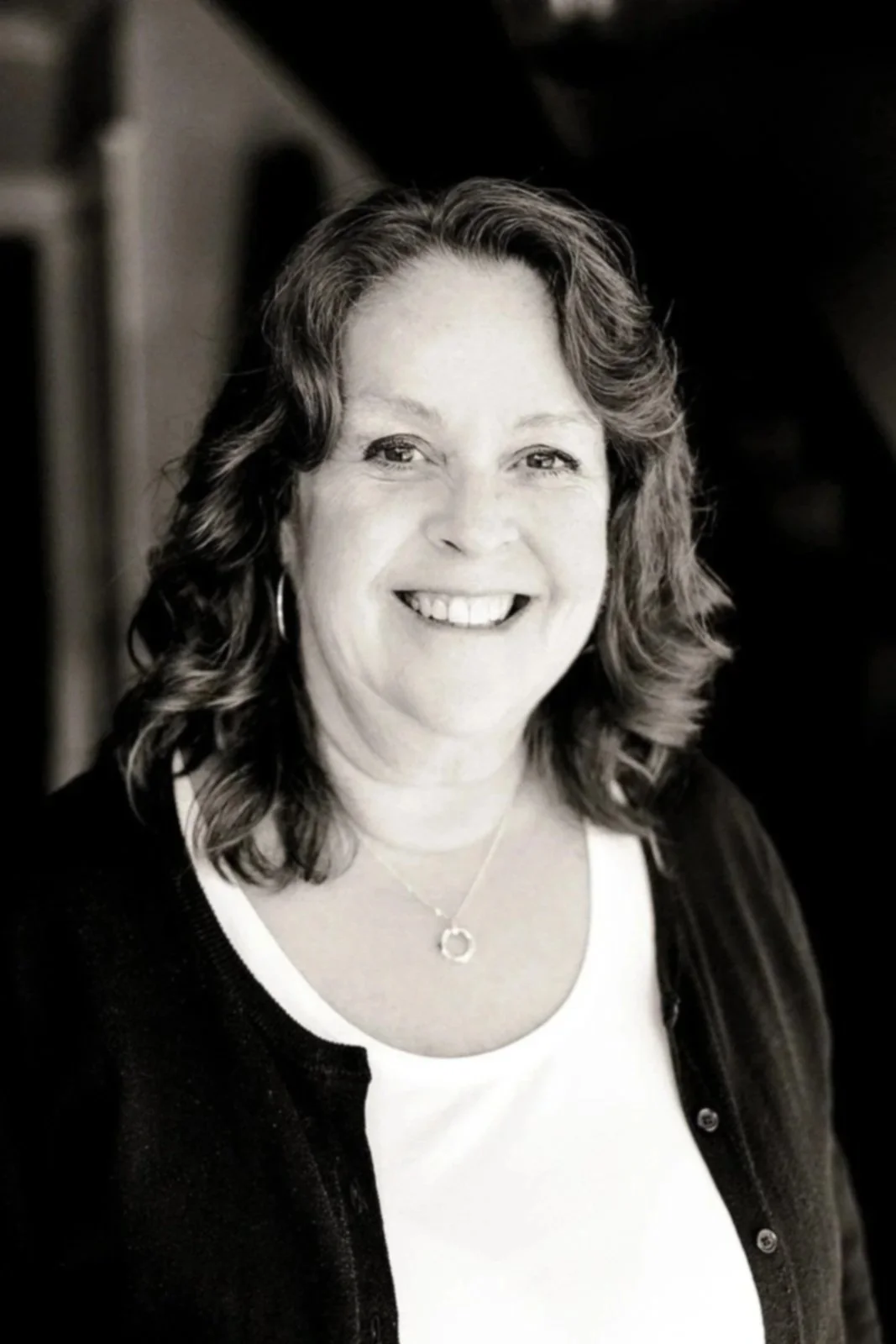 Black and white portrait of a smiling woman with shoulder-length wavy hair, wearing a necklace and a dark jacket over a white shirt.