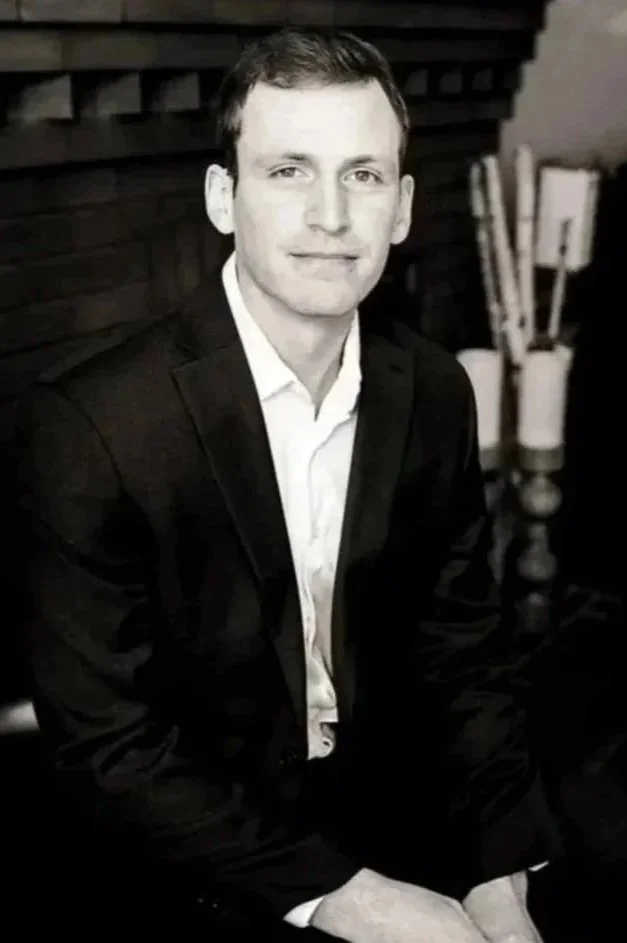 A black-and-white portrait of a man in a suit sitting near a fireplace with lit candles and firewood in the background.