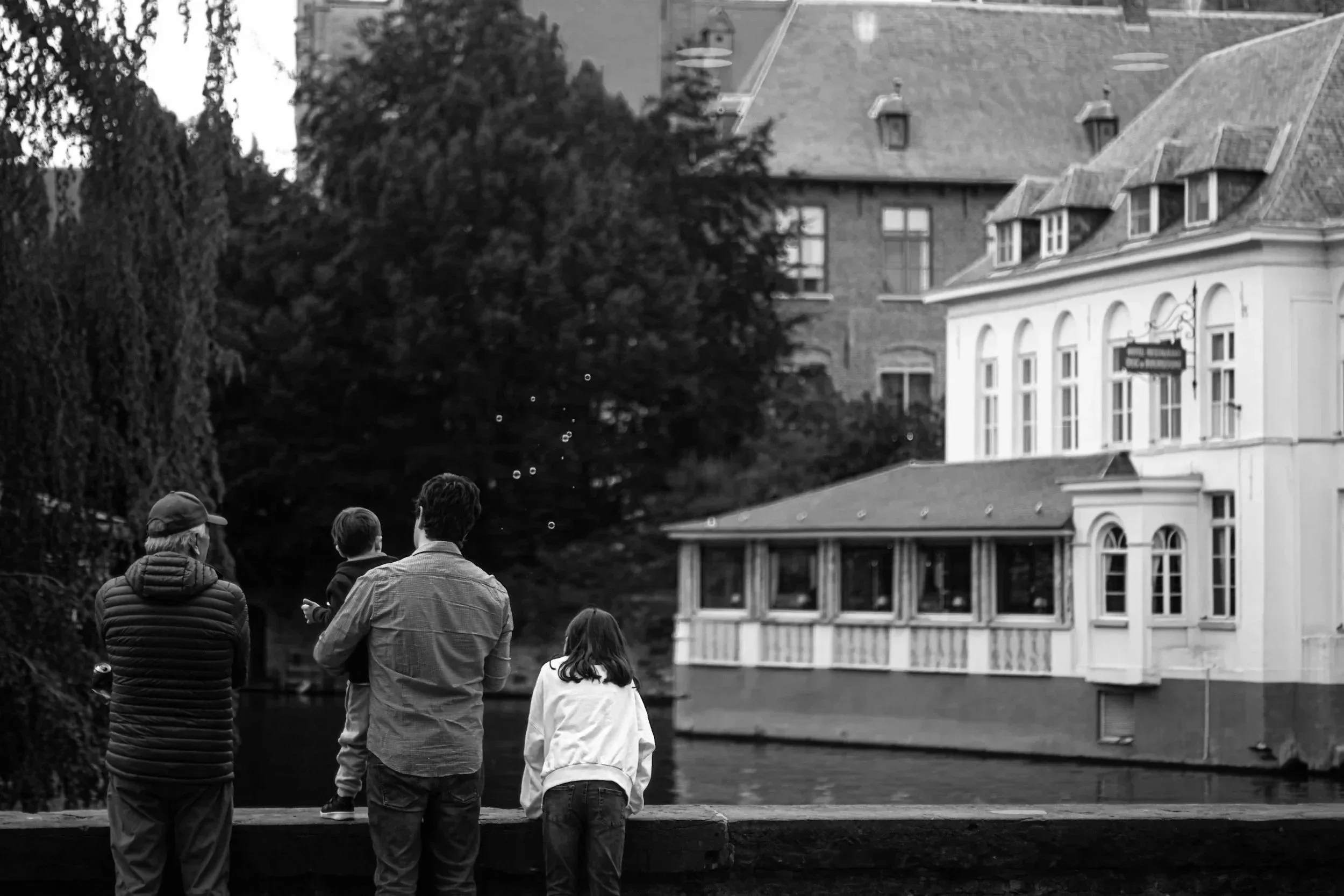 Three people, two adults and one child, stand by a canal in a city with historic buildings and trees, watching bubbles float in the air.