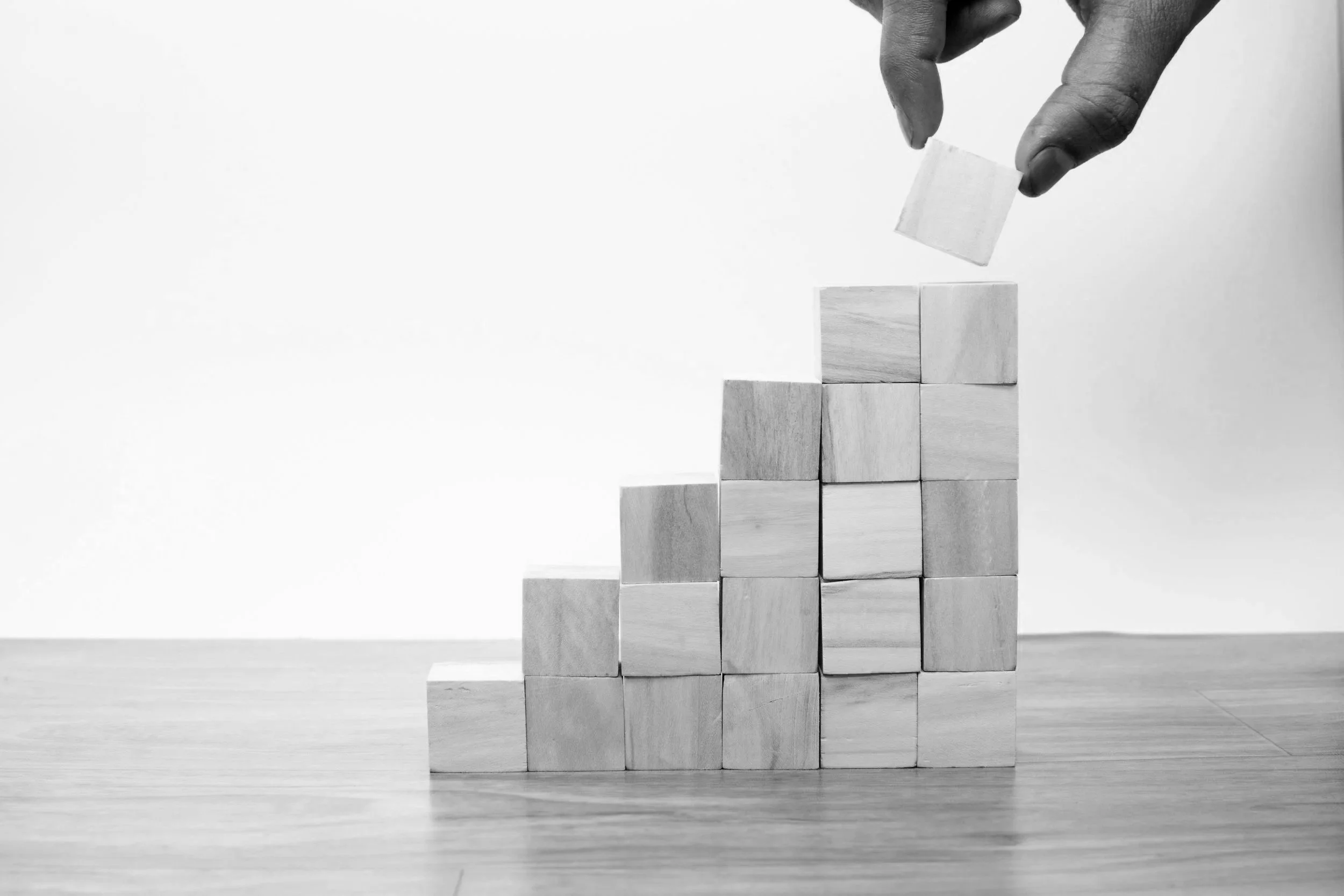 A hand placing a wooden cube onto a stack of similar cubes arranged in increasing height from left to right, resembling a bar chart or growth graph, on a wooden surface with a plain white background.