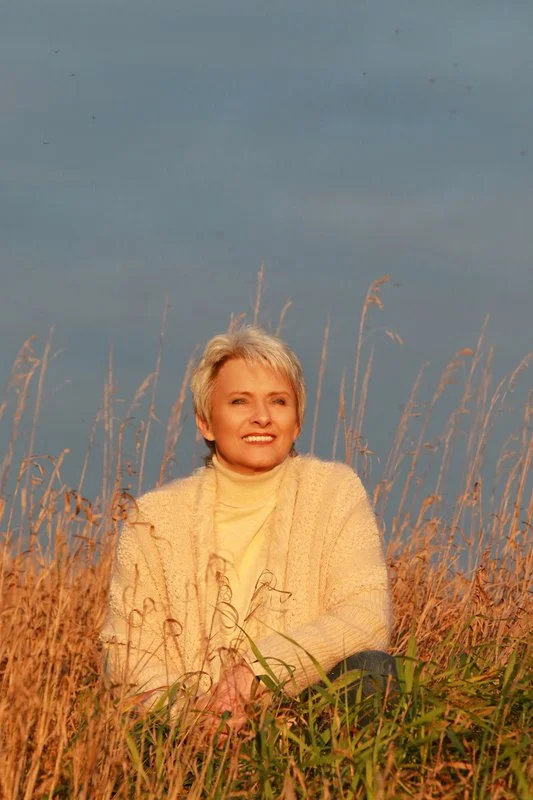 Woman with short blonde hair smiling and sitting among tall dry grass in a field with a blue sky in the background.