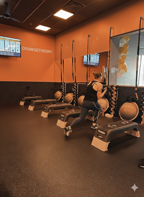 A woman in workout attire using a step platform in a fitness studio with orange walls, exercise balls, resistance bands, and digital screens.