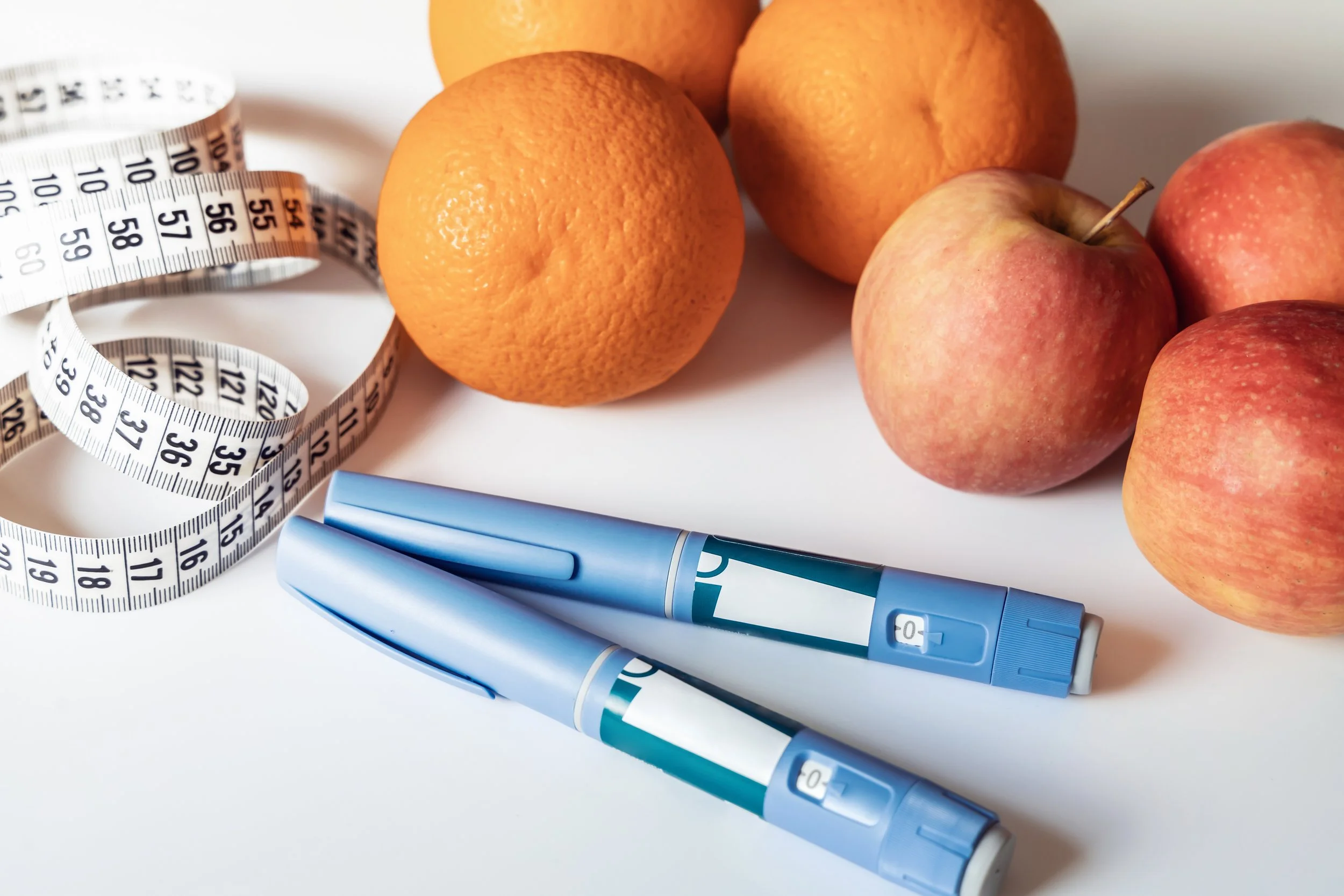 Three oranges and three apples on a white surface, with two blue insulin pens and a white measuring tape nearby.
