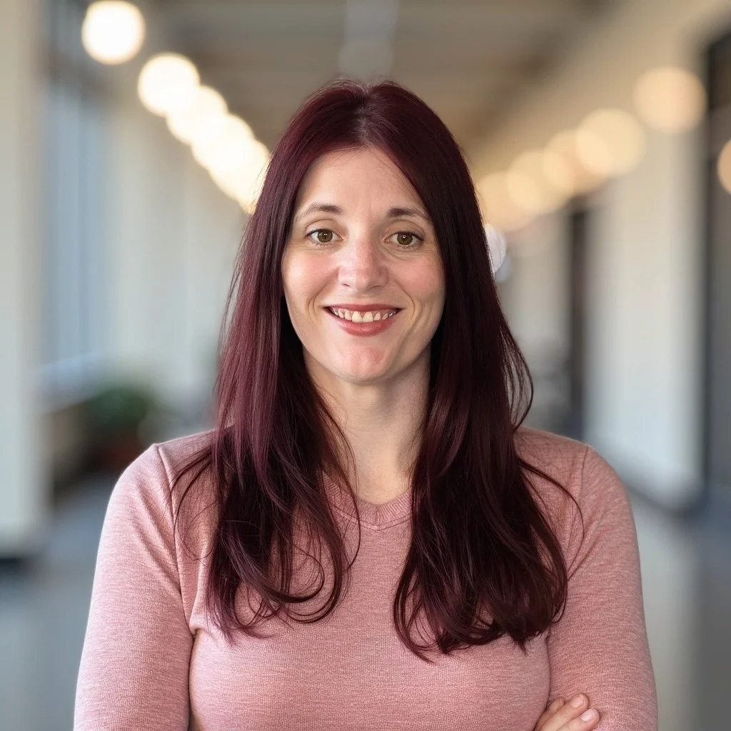 Portrait of a woman with long, dark red hair wearing a pink top, standing in a well-lit corridor with blurred ceiling lights.