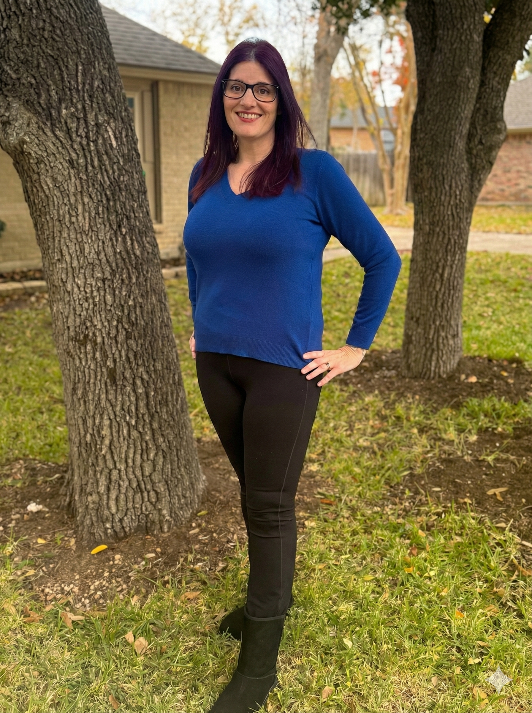 A woman with purple hair, glasses, wearing a blue sweater, black pants, and black boots, standing outdoors among trees with a house in the background.