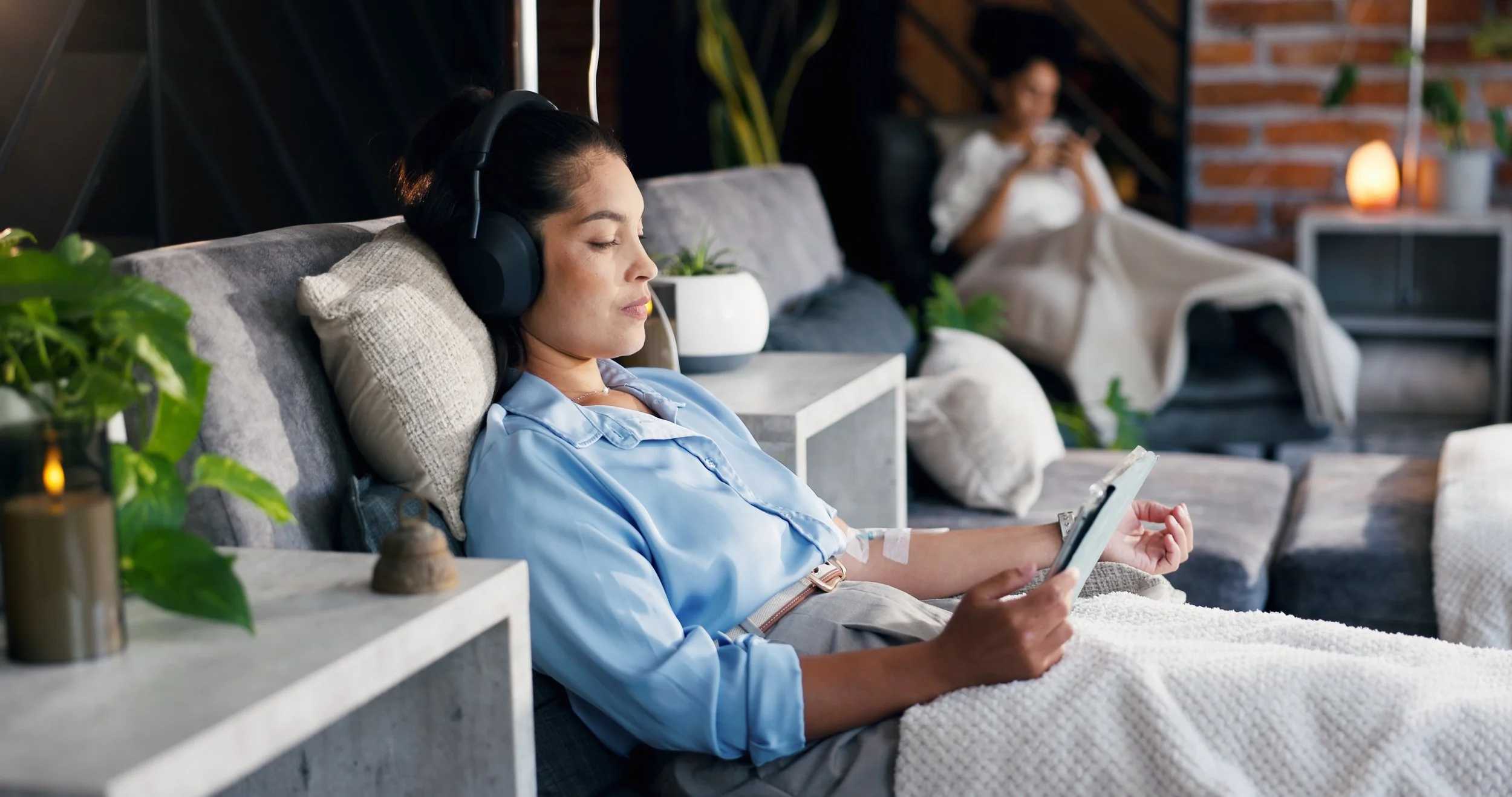 Woman relaxes on sofa wearing headphones and using phone, with another person in the background on sofa.