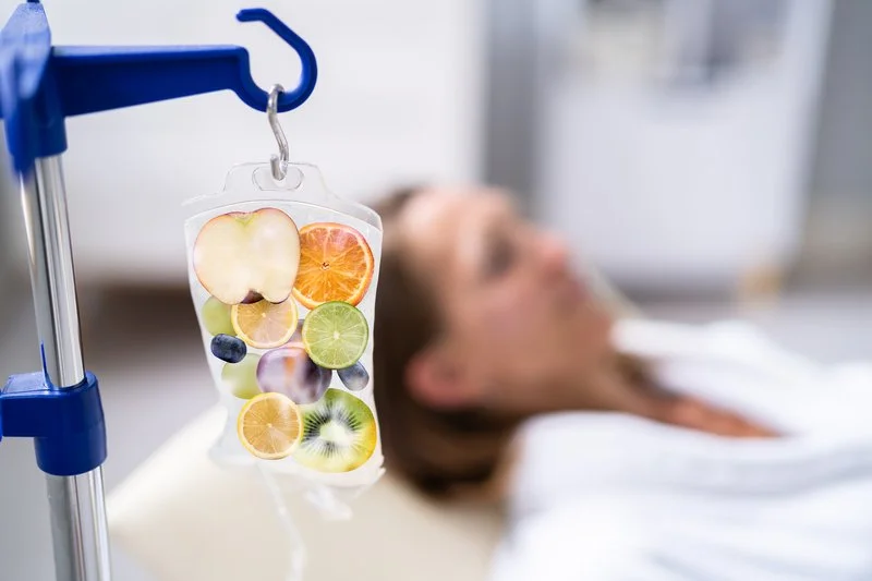 Hospital IV drip bag filled with slices of various fruits, including apple, orange, lemon, lime, blueberry, and kiwi, with a woman lying on a hospital bed in the background.