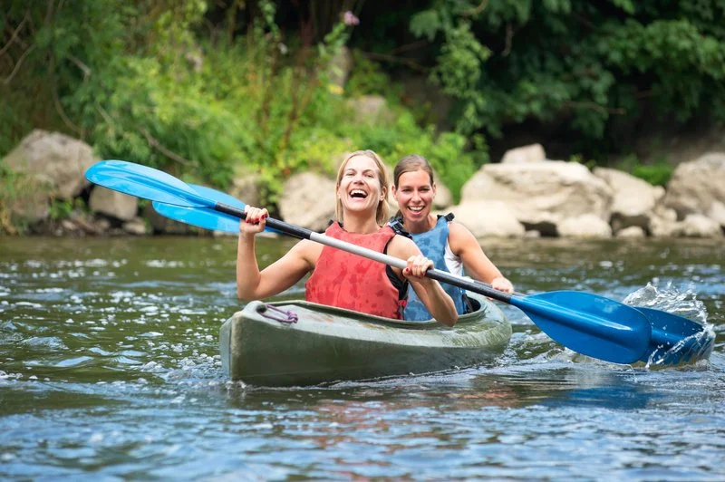 Two women in a kayak paddling on a river surrounded by green trees and rocks, smiling and enjoying their time.
