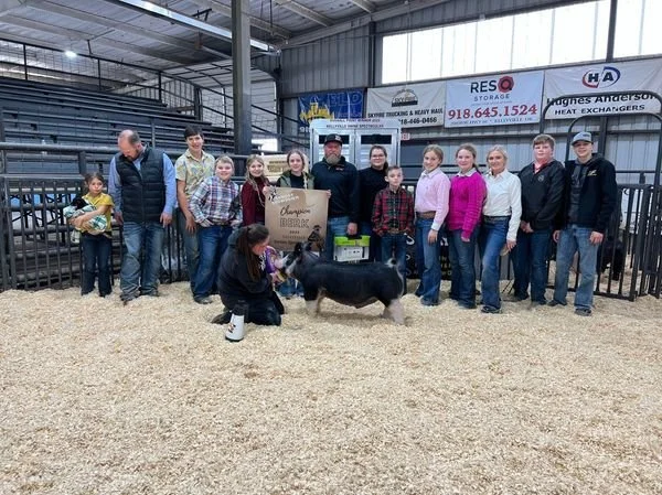 Group of people, including children and adults, standing inside an indoor livestock facility with a small pig, some holding purple ribbons, and a woman kneeling beside the pig.