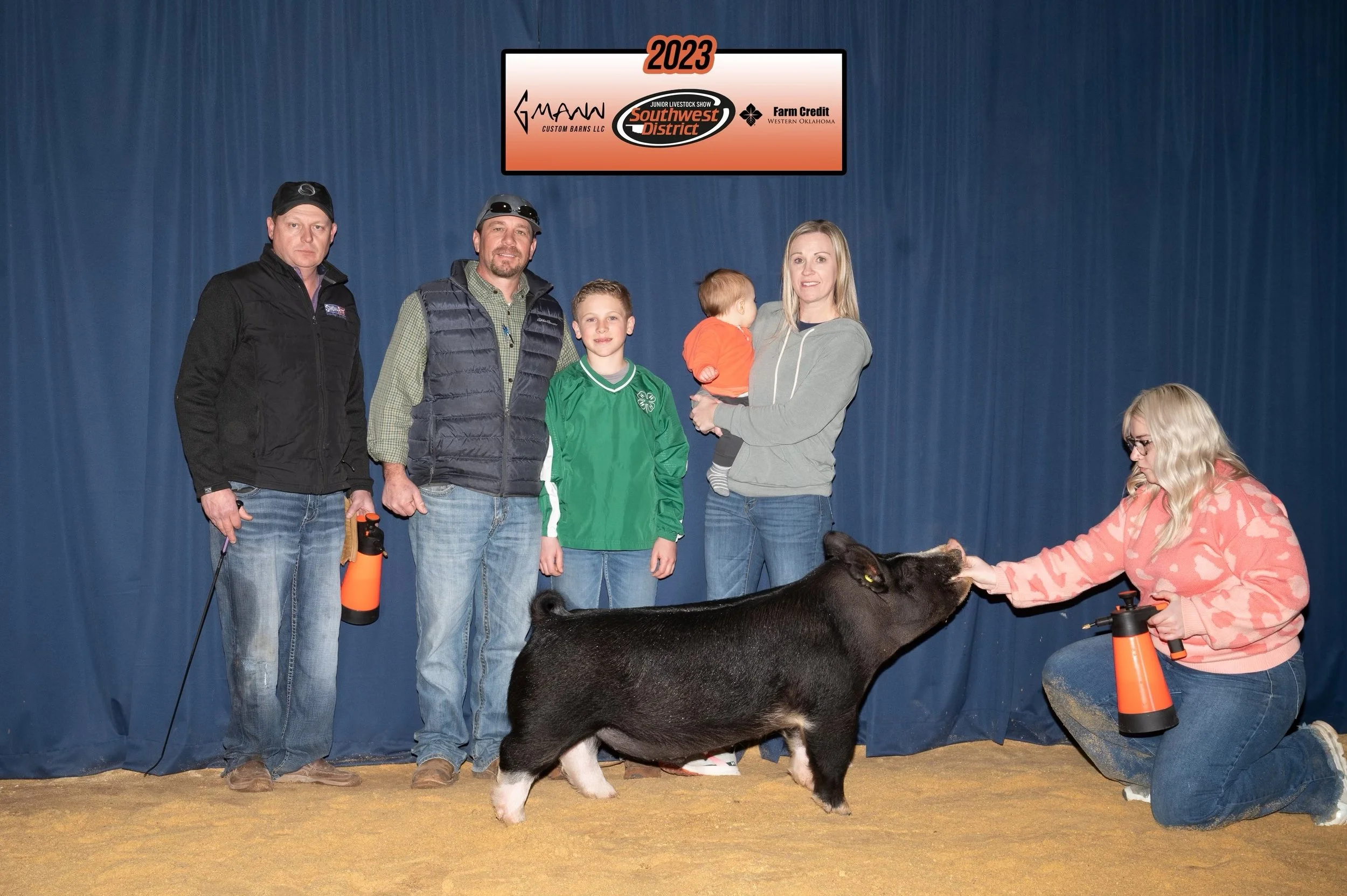 Group of five people and a pig on stage at the 2023 Southwest District Junior Livestock Show, Oklahoma. The stage has a blue curtain, and a sign with event info is above. The woman kneeling on the right is petting the pig, which is being held by a woman standing on the right, holding the pig's snout. The group includes two adult men, a teenage boy, a woman holding a small child, and a smiling pig.