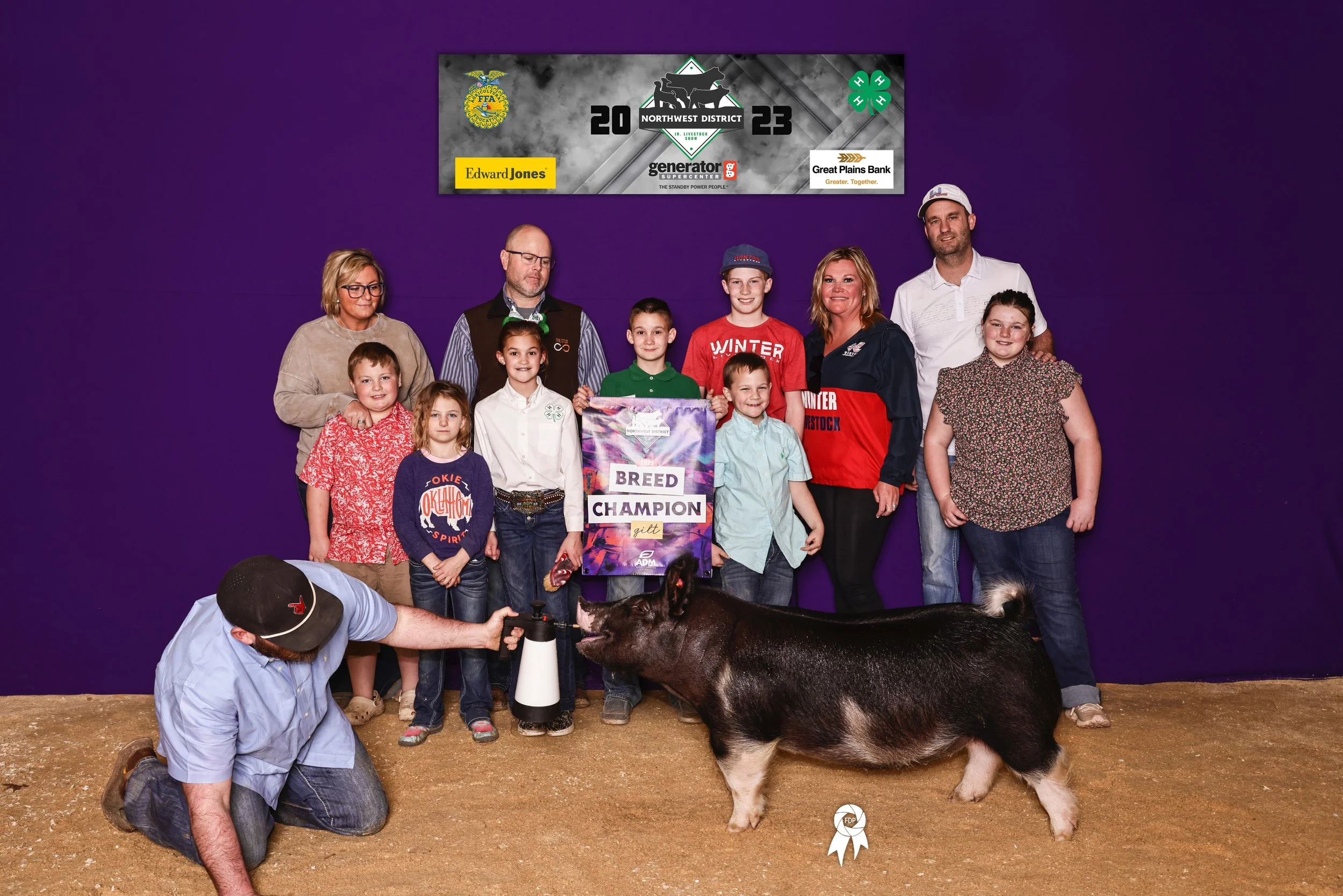 A group of children and adults posing indoors against a purple wall, holding a banner that reads 'Breed Champion'. A man kneels on the ground handing a hand-held device to a pig, which is a black and white pig with a ribbon on the ground nearby. There is a sign above them with various sponsor logos and the year 2023.