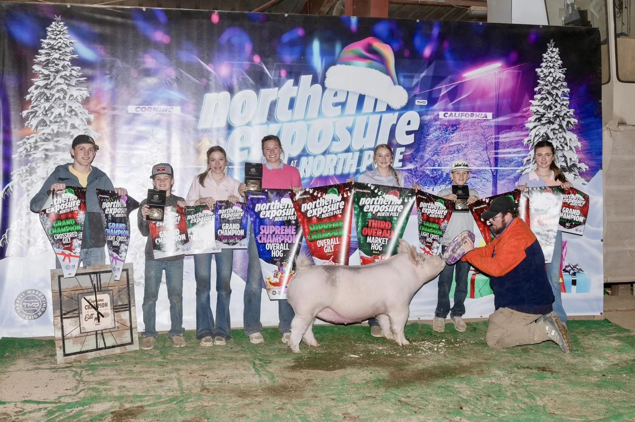 Group of children standing on a stage holding large trophies and awards after a pig farming competition. A farmer is feeding a pig in front of the children. The backdrop displays the event name 'Northern Exposure at North Pole' with Christmas-themed decorations and snowy trees.