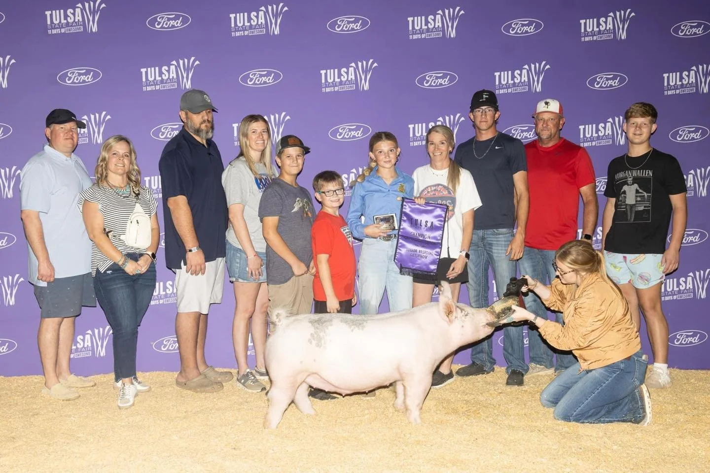 A group of people posing on a purple backdrop with 'Tulsa State Fair' and 'Ford' logos, with a pig in front, and a woman kneeling beside it holding it by the snout.