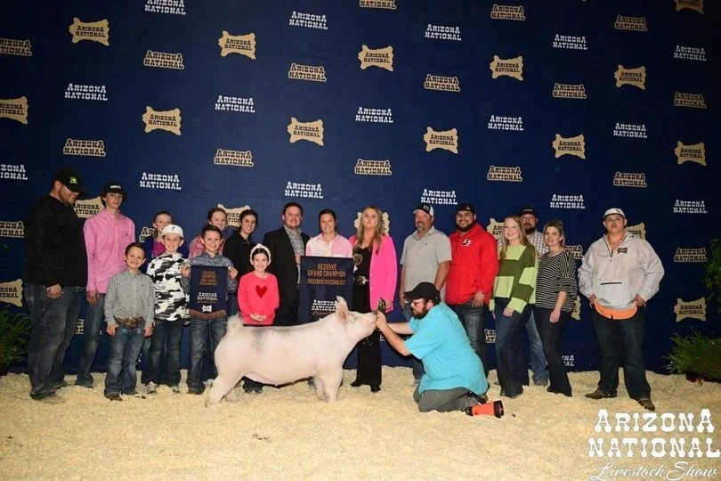 A group of people and children standing on a stage, holding awards, with a pig and a man in a blue shirt kneeling in front of it, at the Arizona National Livestock Show.