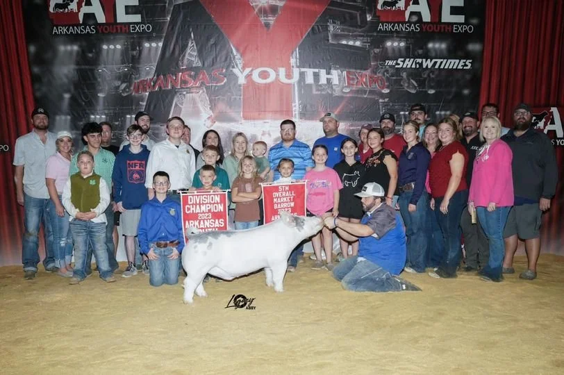 Group of children and adults at Arkansas Youth Expo with a white pig, two people holding the pig's head, and awards in front of a red and black Arkansas Youth Expo banner.
