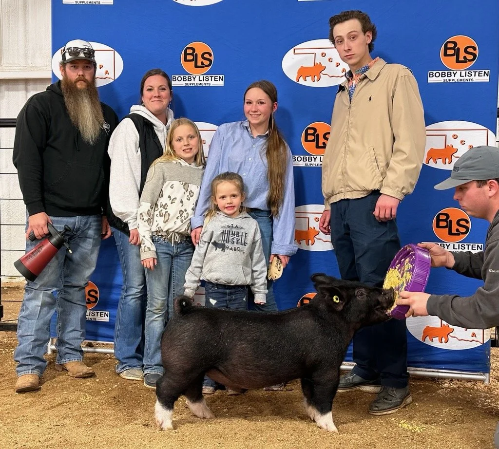 Group of six people, three children, and three adults, standing on dirt ground in front of a blue backdrop with 'Bobby Listen Supplements' logo, a pig illustration, and other graphics. One person is feeding a black piglet with a purple container, while others pose for the photo.