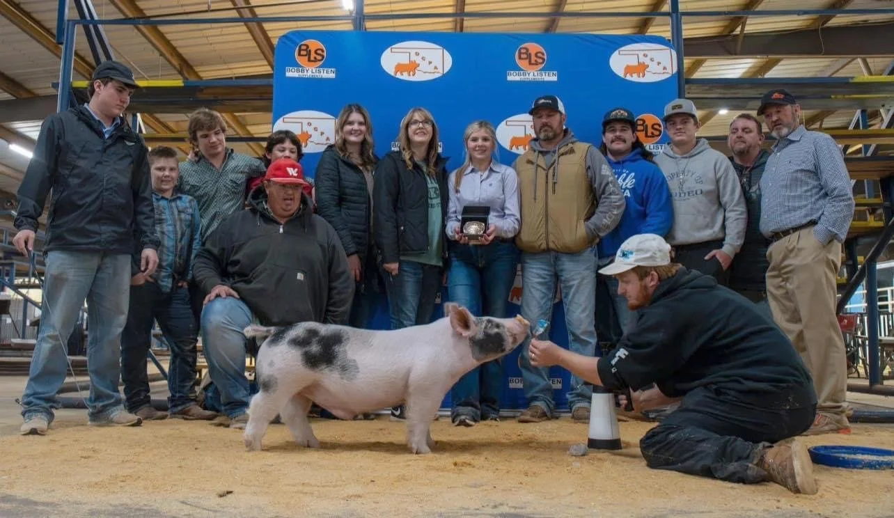 Group of people gathered around a pig at a livestock auction or fair, indoors, with a man kneeling and giving the pig water.