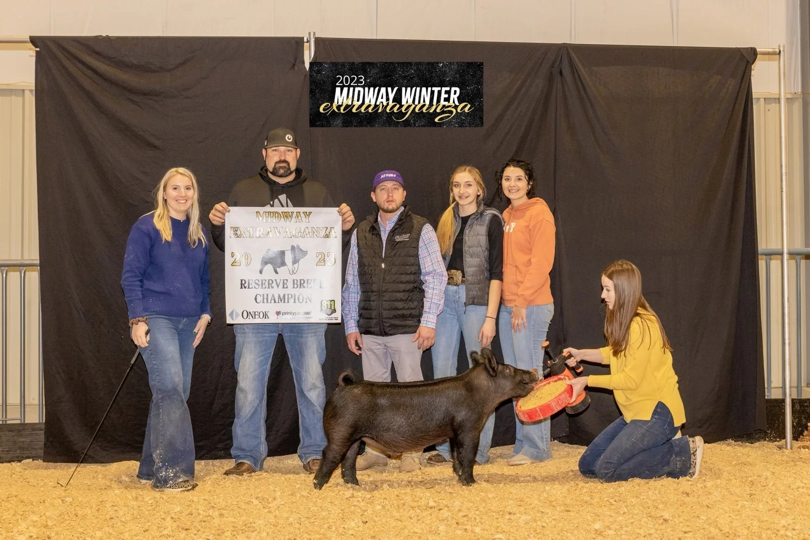 Group of six people, including a woman kneeling with a pig, holding a feeder, at an indoor livestock event with a black backdrop, celebrating the 2023 Midway Winter Extravaganza Reserve Breed Champion, with a sign and pig in front of them.