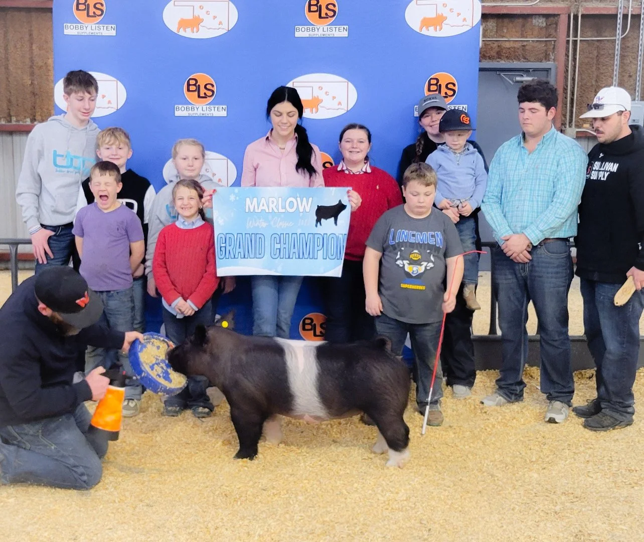 A group of children and adults standing around a pig that is being awarded a blue ribbon at a fair or livestock event. One girl holds a sign that says 'Marlow 울트라 Grand Champion.'