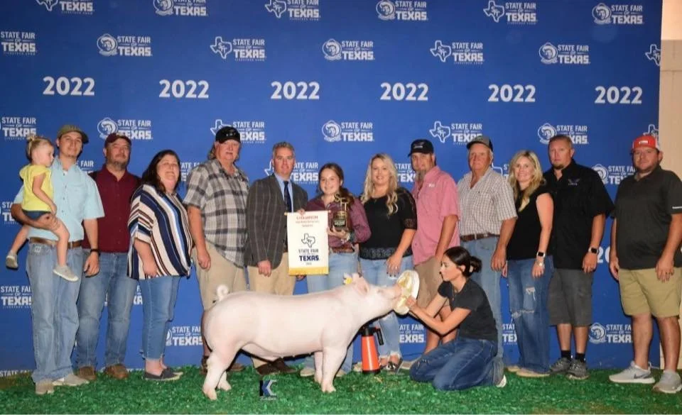 A group of people at the Texas State Fair 2022, posing with a pig and a trophy. One woman is feeding the pig, which is standing on grass in front of a blue backdrop. The backdrop features the Texas State Fair logo and the year 2022 multiple times.