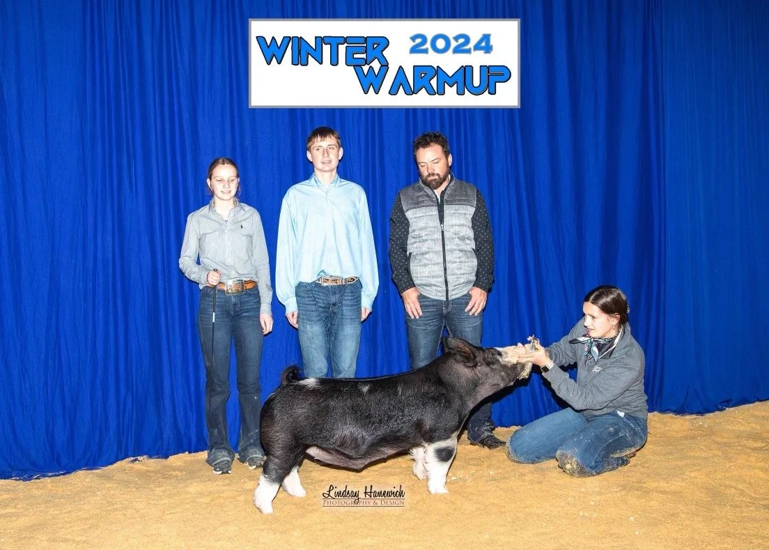 Group of three young people and an adult with a pig on a dirt floor, posing in front of a blue curtain with a sign that says "Winter Warmup 2024" at an indoor event.