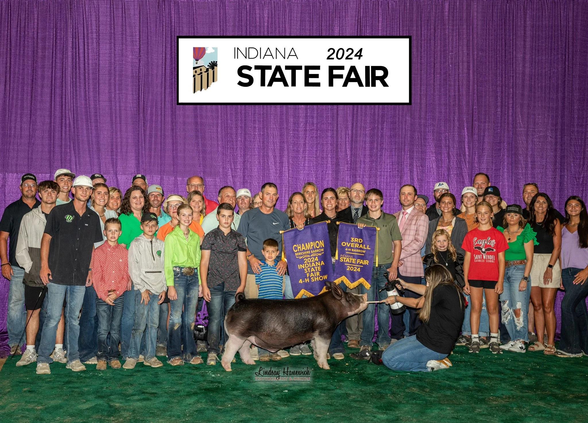 Group of people at the Indiana 2024 State Fair on a stage with purple backdrop, holding banners for champion and third overall winners in the 4-H show, with a pig in front and a photographer kneeling.