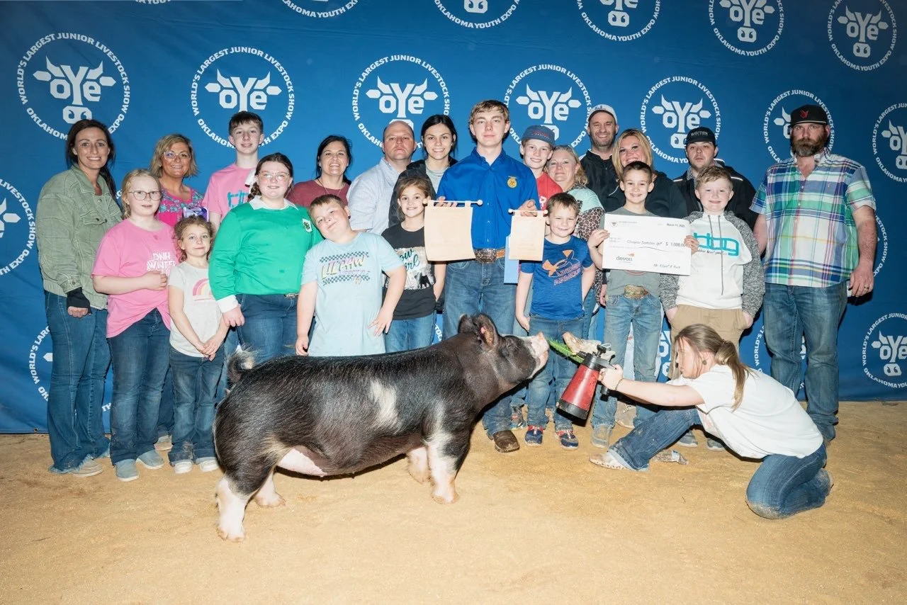 Group of children and adults at Oklahoma Youth Expo, standing behind a pig and a young girl holding a fire extinguisher. The group appears to be celebrating, with some holding a large check and awards, in front of a blue backdrop with the event logo.