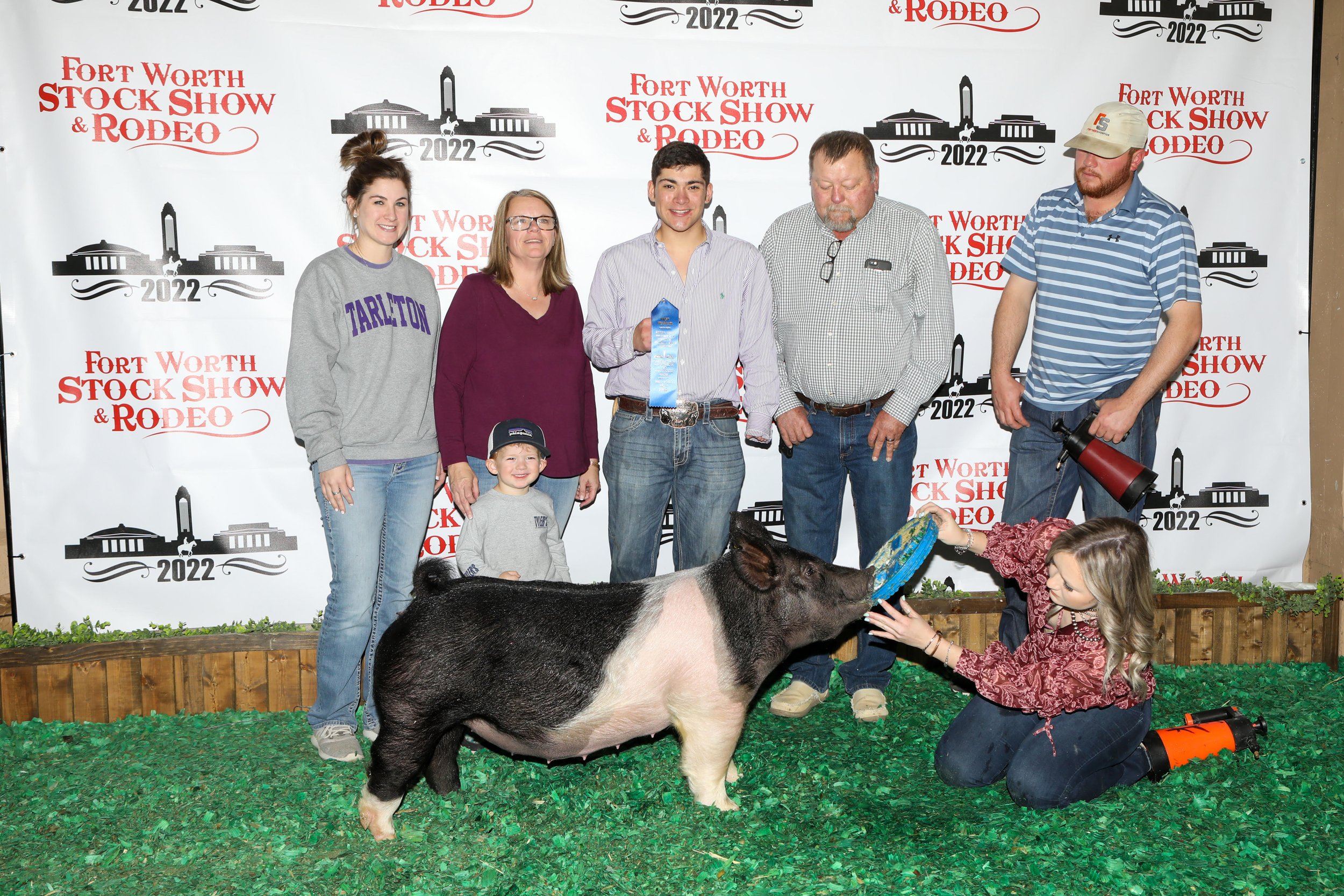 Group of six people, including a young boy, standing behind a pig, with a woman kneeling in front holding a frisbee near the pig's mouth. The background displays a step and repeat banner with the event name 'Fort Worth Stock Show & Rodeo 2022'.