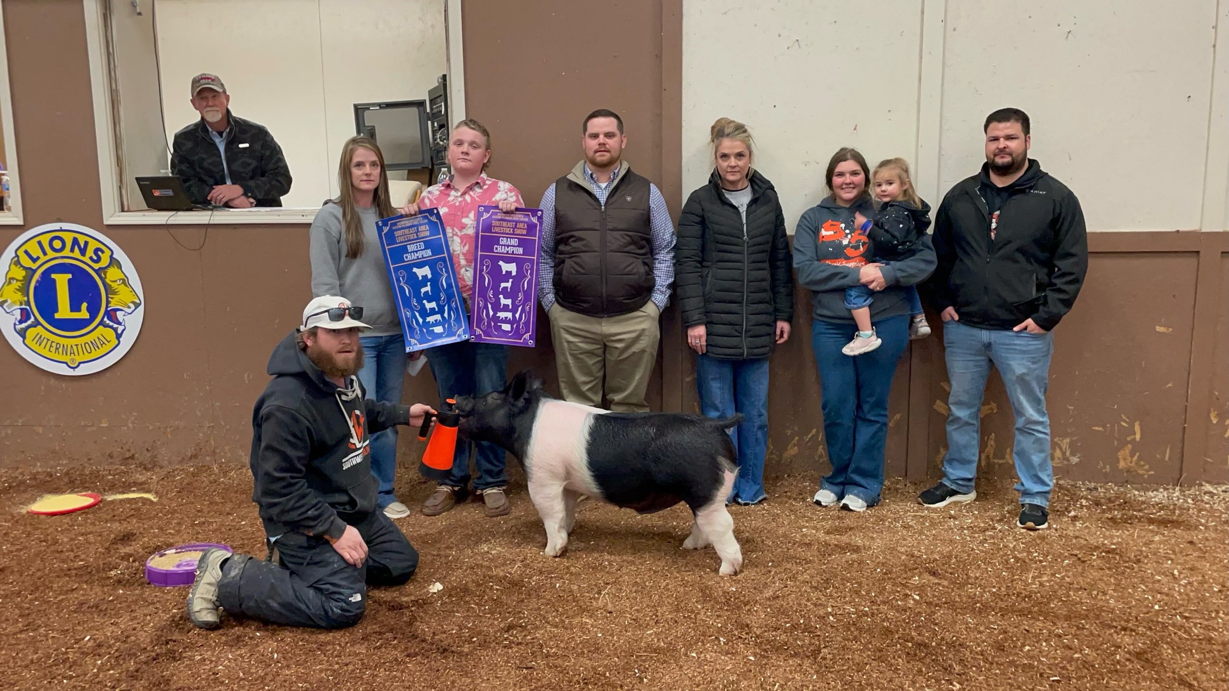 A group of people, including children and adults, standing behind a small pig at an indoor livestock show. The pig is Black and White, and the group appears to be celebrating as some hold large blue ribbons that signify grand and breed champion awards. Someone kneels in front of the pig, holding it, with a barn or livestock arena setting and a Lions International logo visible on a sign to the side.
