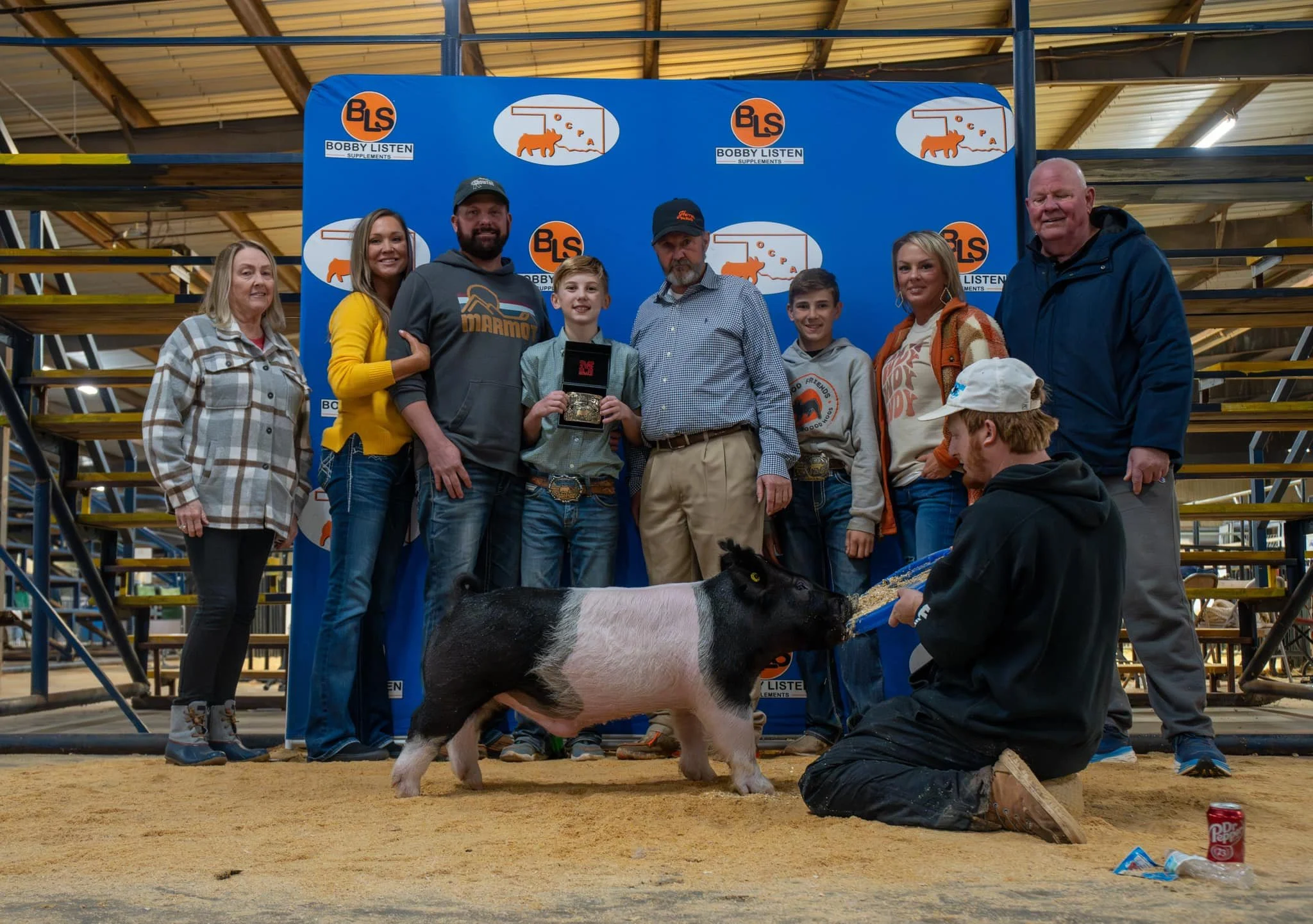 Group of people standing in front of a blue backdrop with logos, with a pig and a man with a butterfly knife in front of them, inside a barn.