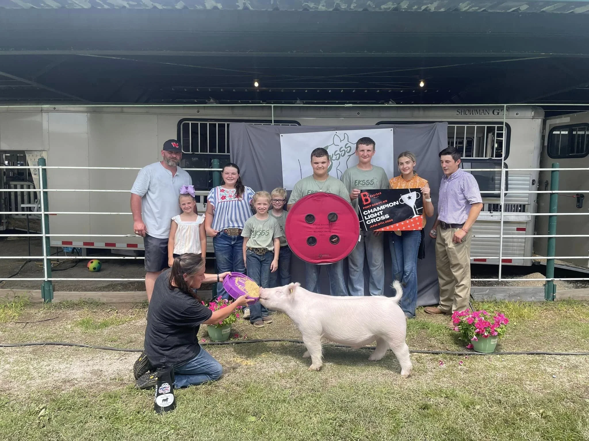 Group of people, including children and adults, standing inside a metal-fenced area with a pig and a woman kneeling in front, feeding the pig. The background features a trailer and a banner. There are pink flower pots on either side.