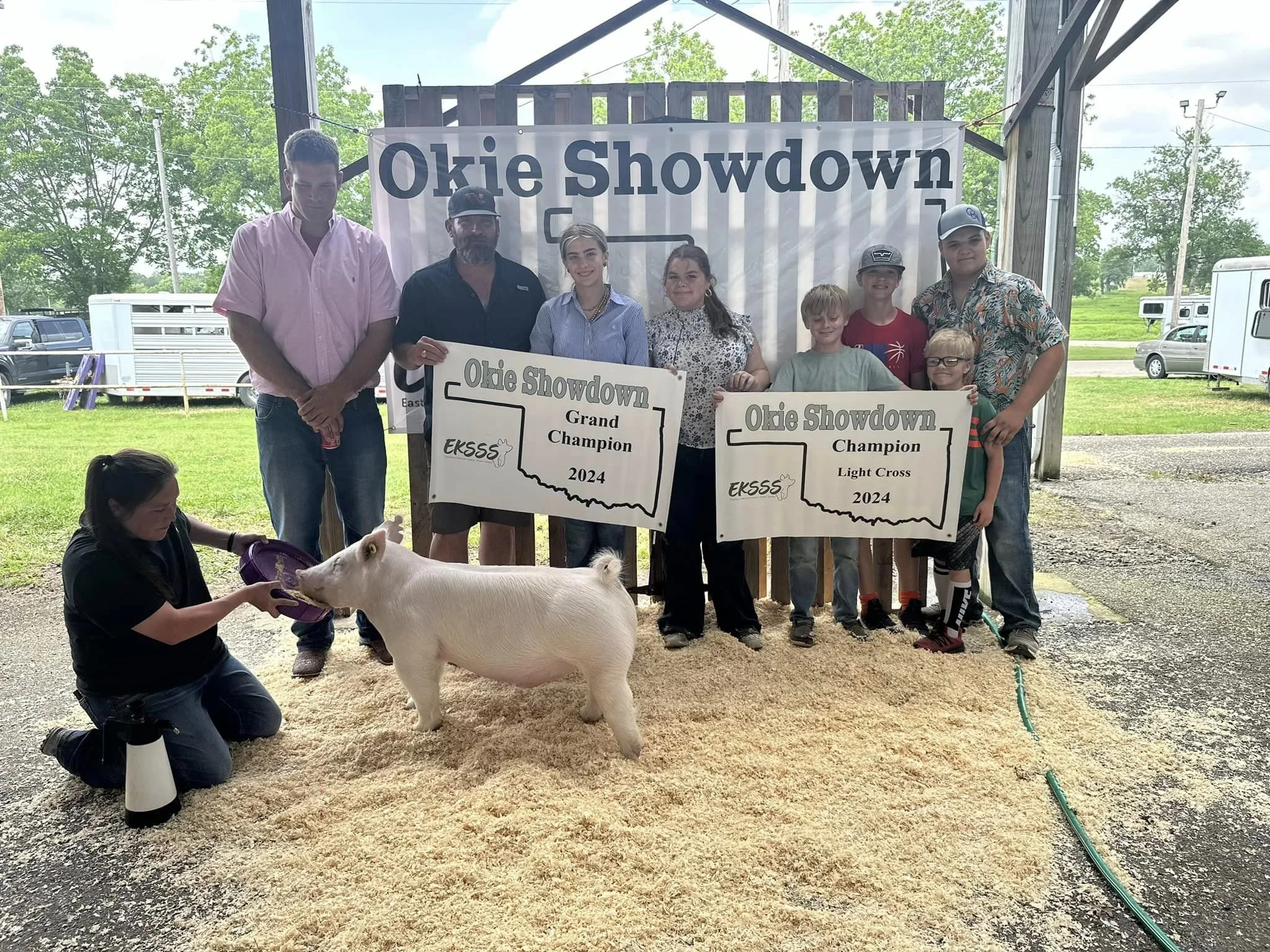 Group of people posing with trophies and ribbons, holding signs that read "Okie Showdown" and "Grand Champion 2024" and "Light Cross 2024", with a pig in the foreground, inside a barn or covered area, with green trees and vehicles outside.