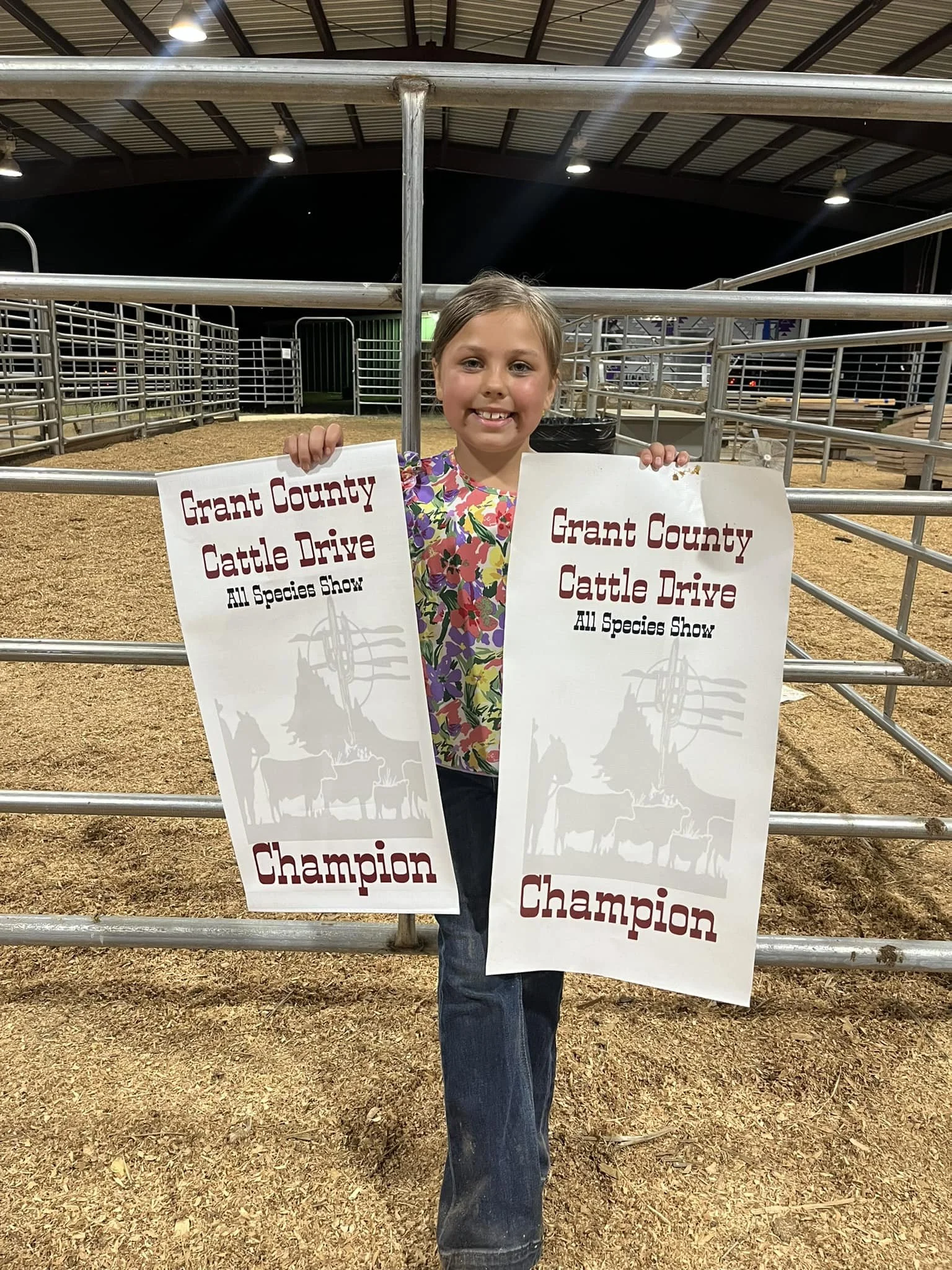 A young girl smiling and holding two signs that read "Grant County Cattle Drive All Species Show Champion". She is standing inside a livestock pen at an indoor event, with metal fencing and a dirt floor, under a large roof with lights.