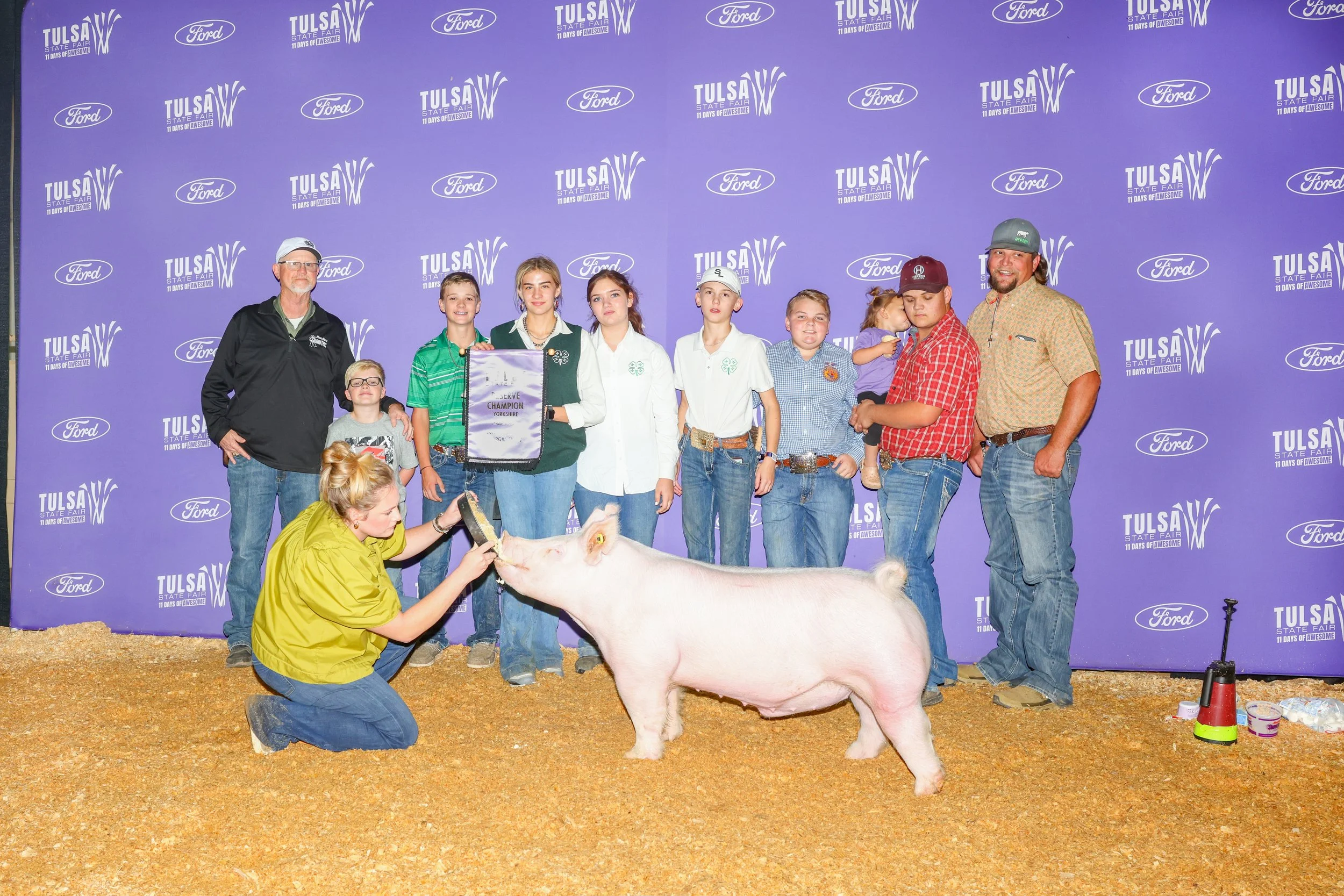 A group of people, including children and adults, standing behind a pink pig at a livestock event, with a woman kneeling and feeding the pig on the ground. The background features a purple wall with logos for Tulsa State Fair and Ford.