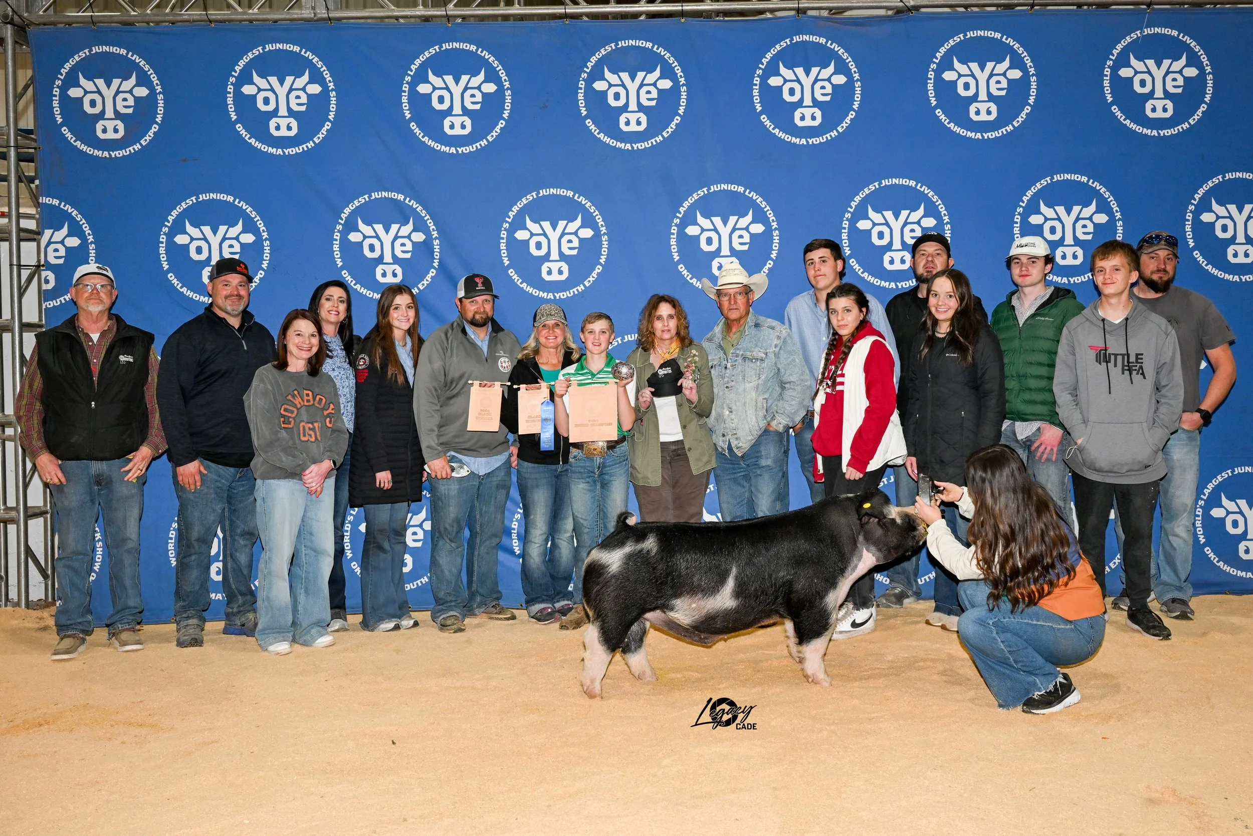 Group of people posing with a pig at the Oklahoma Youth Expo, Oklahoma Youth Livestock Show, standing on a sandy surface in front of a blue backdrop with logos.