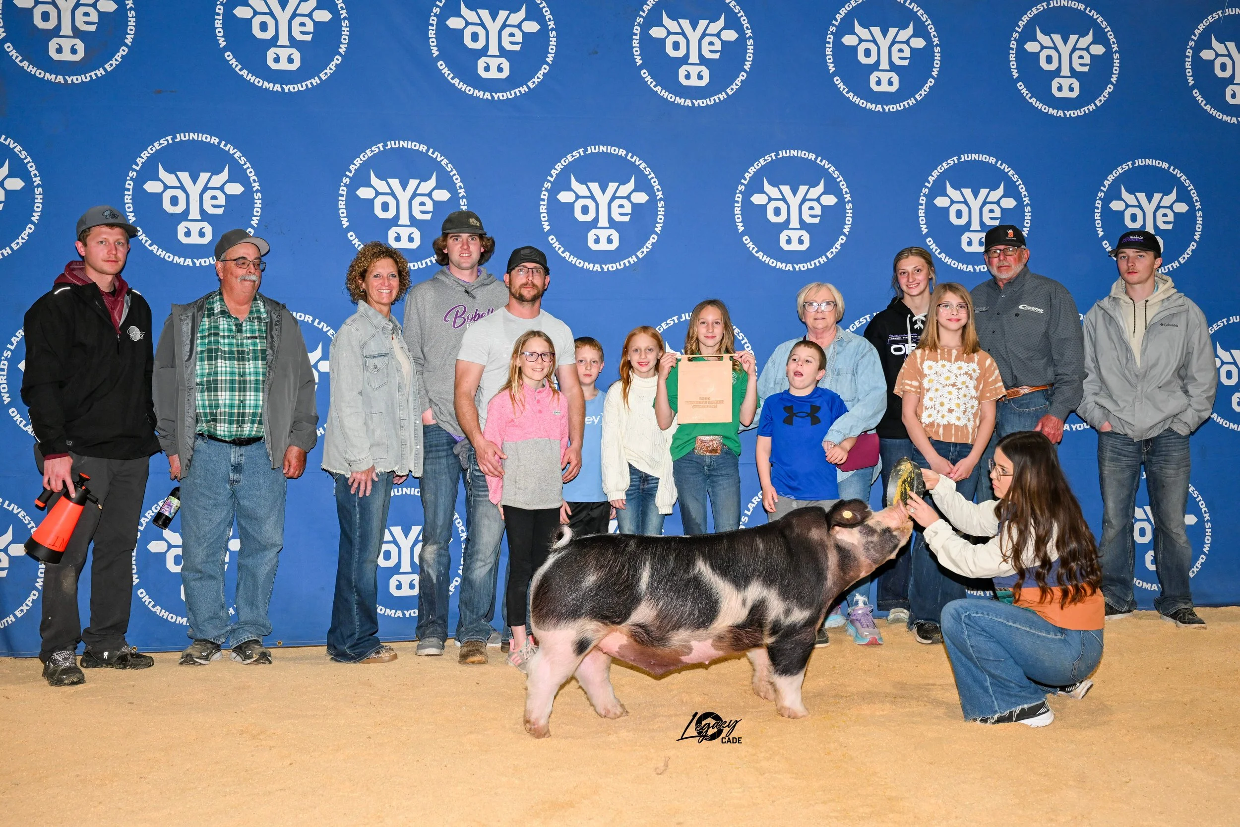Group of people, including children and adults, gathered in front of a blue backdrop with white logos and text that reads 'Oklahoma Youth Expo' and 'World's Largest Junior Livestock Show'. A girl is kneeling and feeding a pig, while others stand behind them, some holding awards or items.