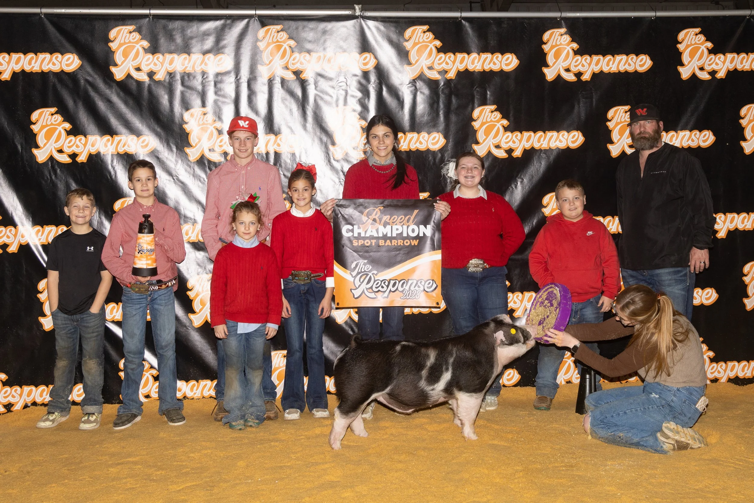 Group of children and adults at a farm event, standing on a yellow floor in front of a black backdrop with orange and white logo of 'The Response,' celebrating a champion spot on a pig named 'Spot Barrow,' with a pig in front and a young woman petting it.