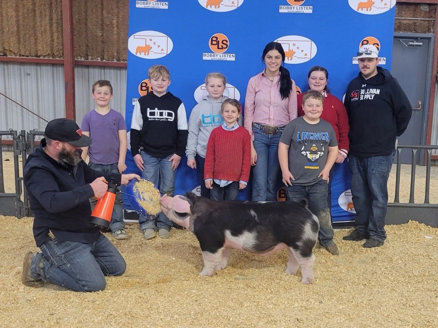 A group of children and two adults posing behind a pig in an indoor farming setting. The man kneeling in front is feeding the pig food from a ladle, and the pig is standing on straw bedding. Behind the group, there is a blue backdrop with farm-related logos and text.