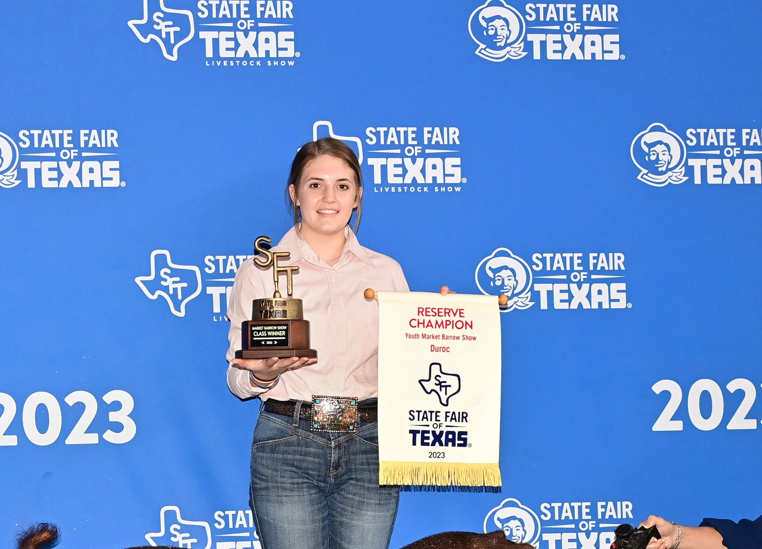 Young woman standing in front of a blue backdrop with 'State Fair of Texas' logos, holding a trophy and a banner that reads 'Reserve Champion' at the Youth Market Barrow Show, 2023.