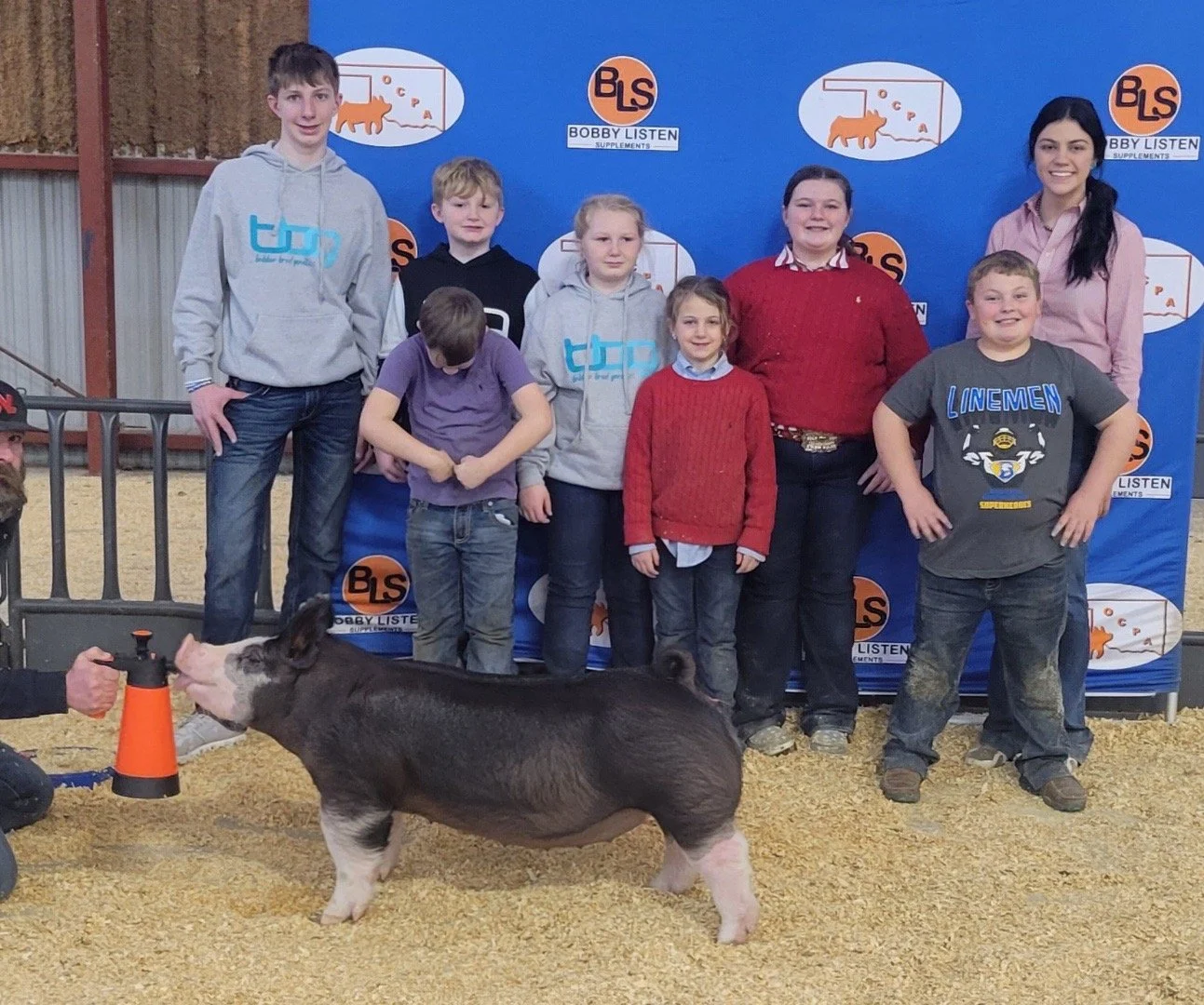 Group of children and adults posing in front of a blue backdrop with logos at a livestock event, with a pig being shown and a handler holding a cone