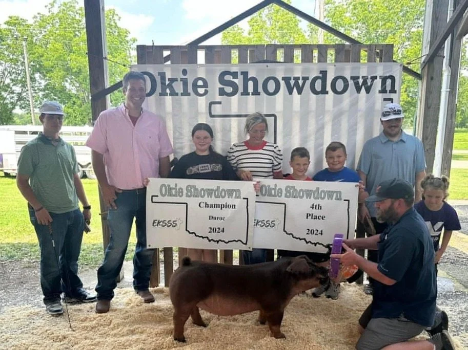 Group of people at an Ohio Showdown event with a kid winning with a pig, holding large checks for champion and 4th place, outdoors with a wooden structure and trees in background.