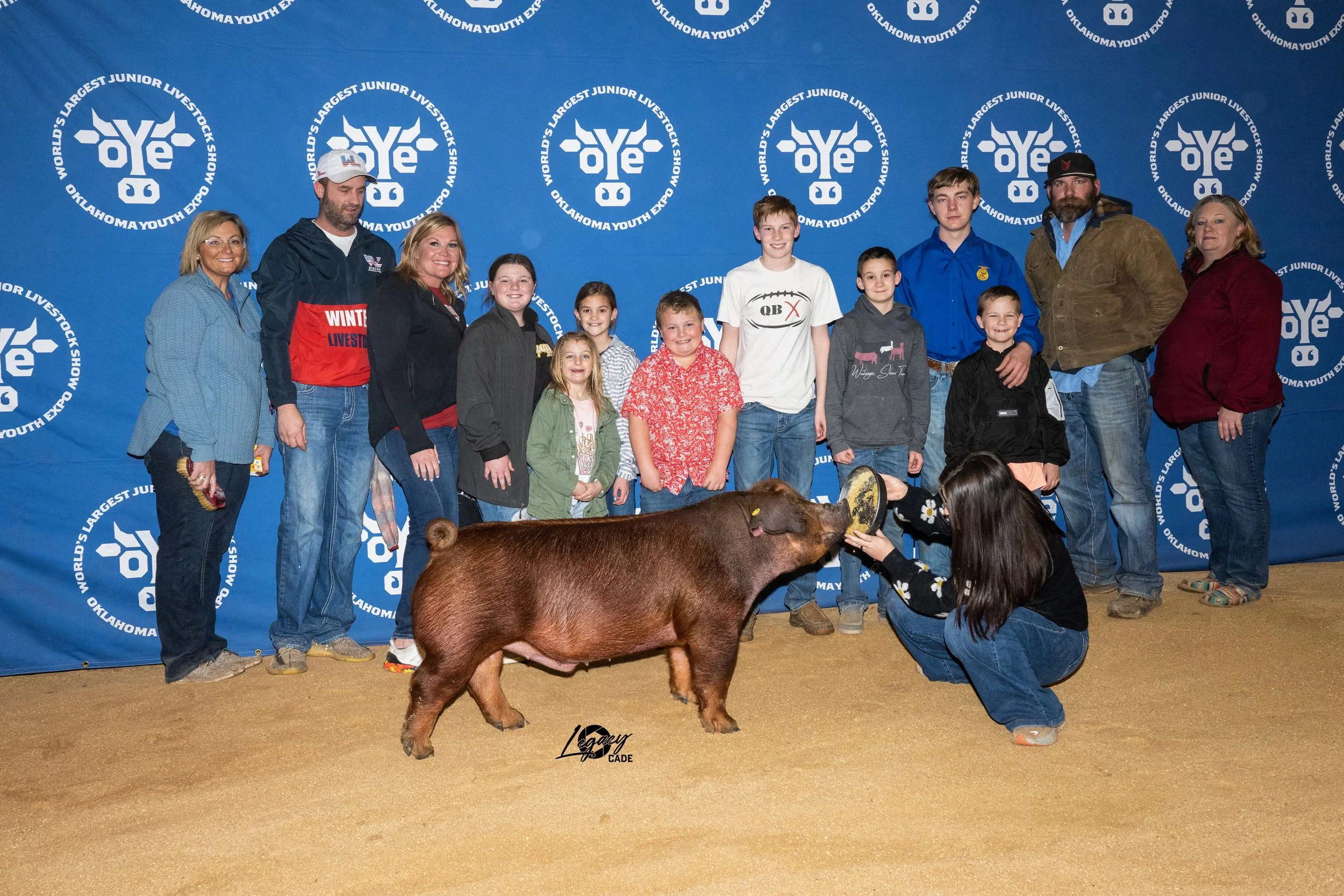 A group of people, including children and adults, standing in front of a blue backdrop with Oklahoma Youth Expo branding. They are gathered around a pig, which is being fed by a girl kneeling in front of it.