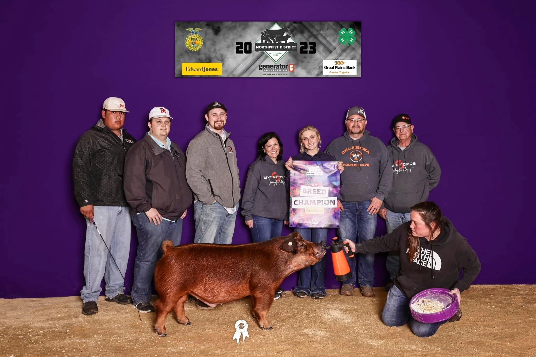 Group of seven people standing behind a pig, which is holding a spray bottle near its face. One woman kneels in front of the group, holding a purple bowl. They are indoors on a dirt floor, with a purple wall behind them. Some of the people wear sweatshirts with 'SWINFORD'S' and 'OKLAHOMA' logos. The woman in the center holds a colorful sign that reads 'Breed Champion.' The top of the image shows a banner with various sponsor logos and text indicating an event for the NW District 2023.