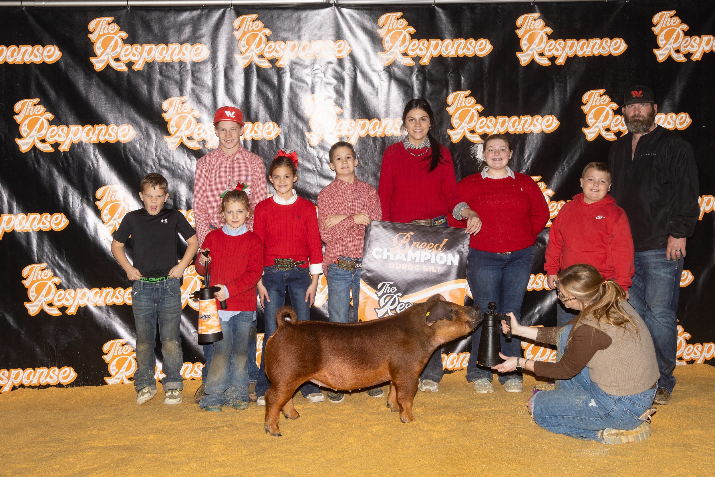Group of children and adults standing on a stage, some holding awards, with a pig in front and a woman holding a spray bottle, during a livestock event.