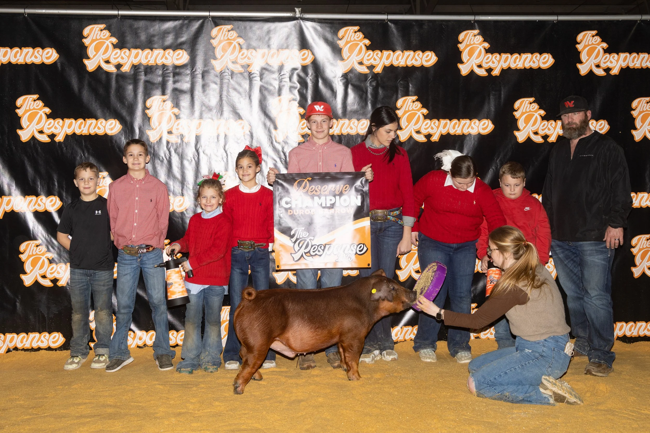 Group of children and adults at a livestock award event with a pig, holding a champion banner, on stage with a black backdrop that says 'The Response' repeatedly.