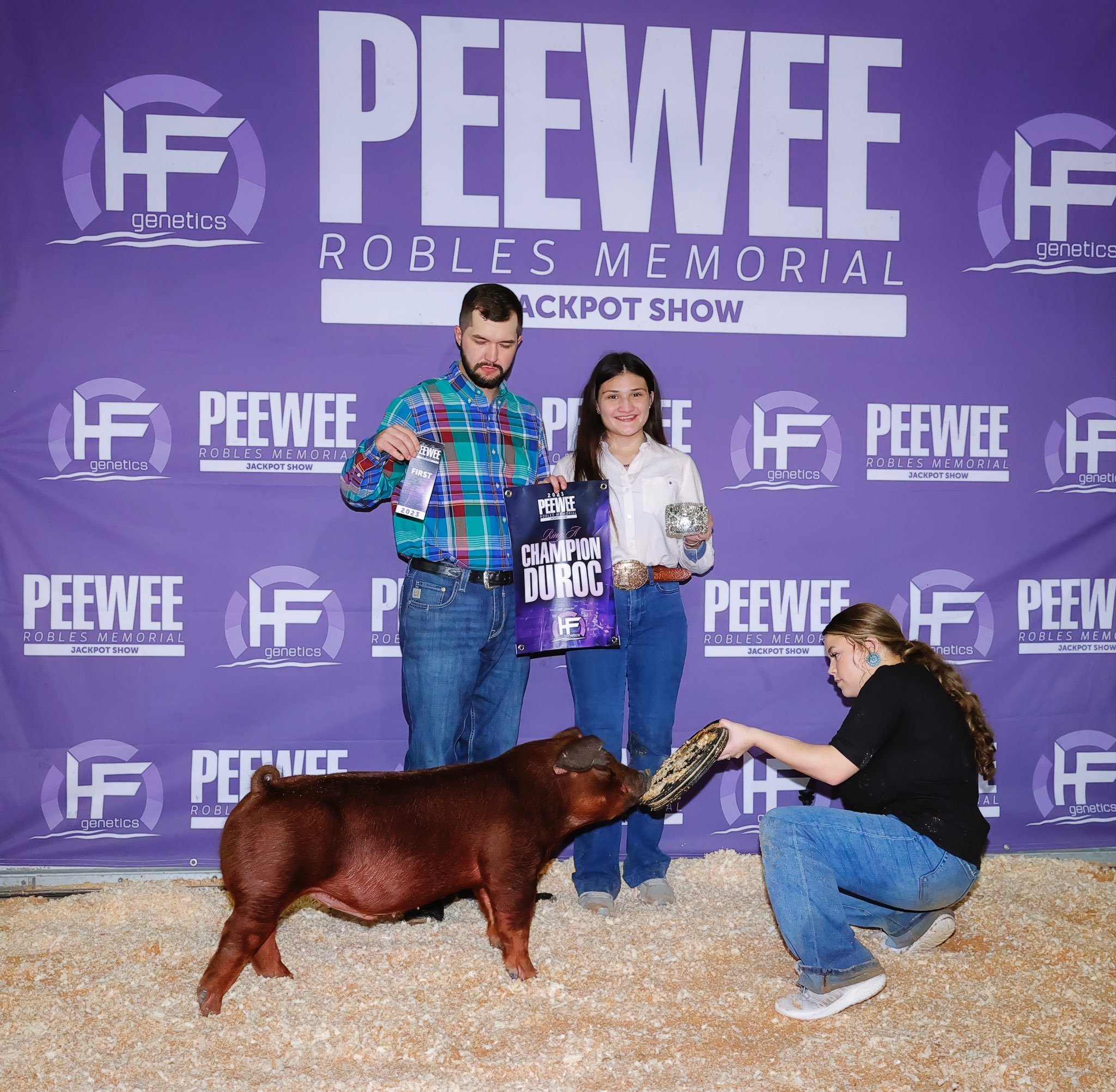Two people on stage at a livestock show, holding awards and a banner that reads 'Champion Duroc,' with a woman kneeling holding a piglet and feeding it.