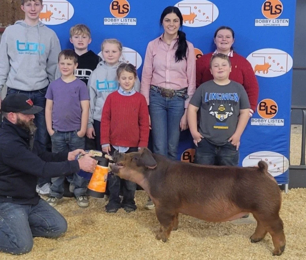 A group of children and two women standing behind a pig at an indoor petting zoo or agricultural event, with a blue backdrop featuring the BLS (Bobby Listen Supplements) logo and an outline of a pig. A man kneels in front handing treats to the pig, which is reaching out to eat.