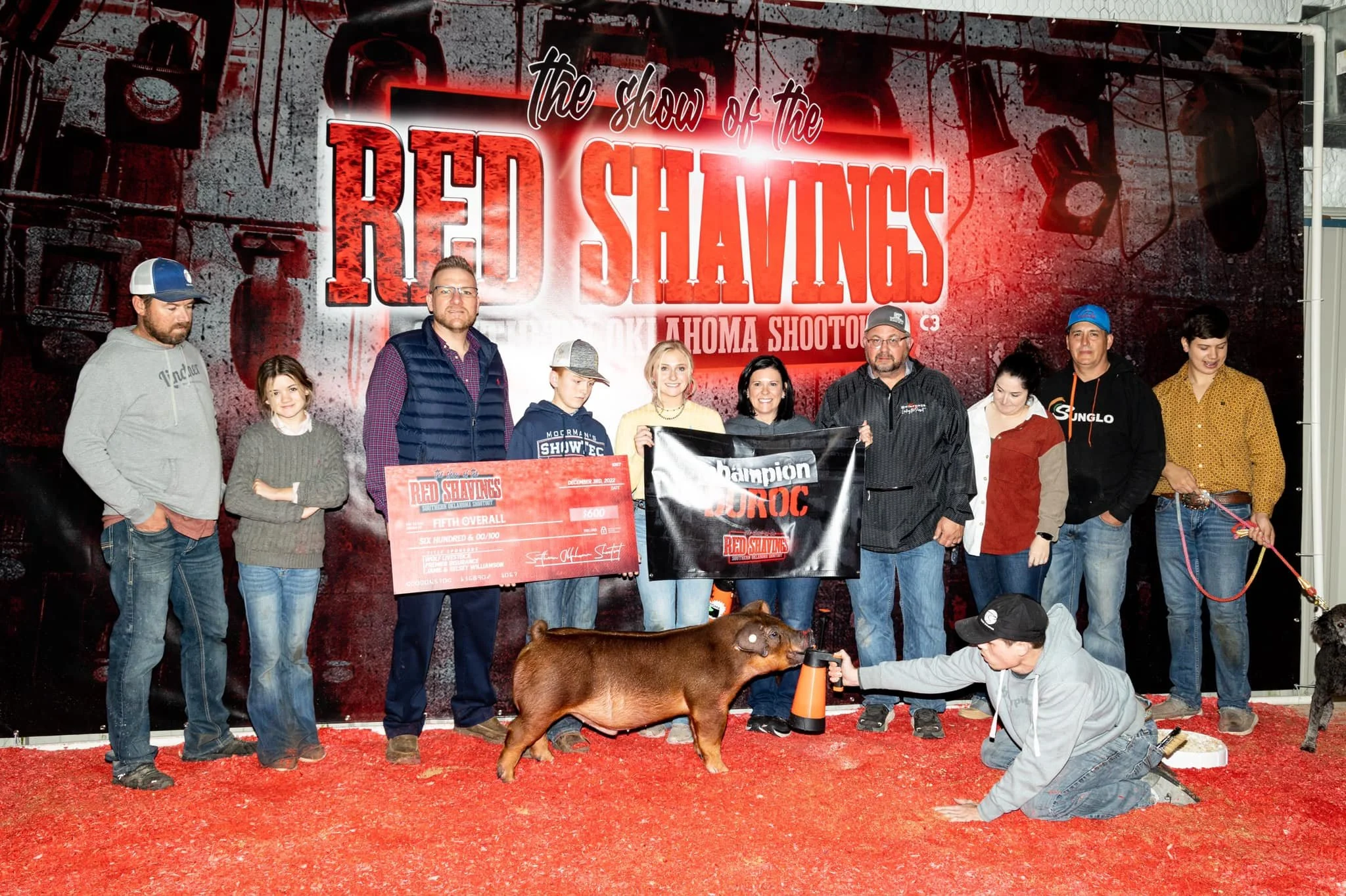 Group of people on stage at the Red Shavings Oklahoma Shootoff, with a pig and a person dressed as a judge in the foreground.