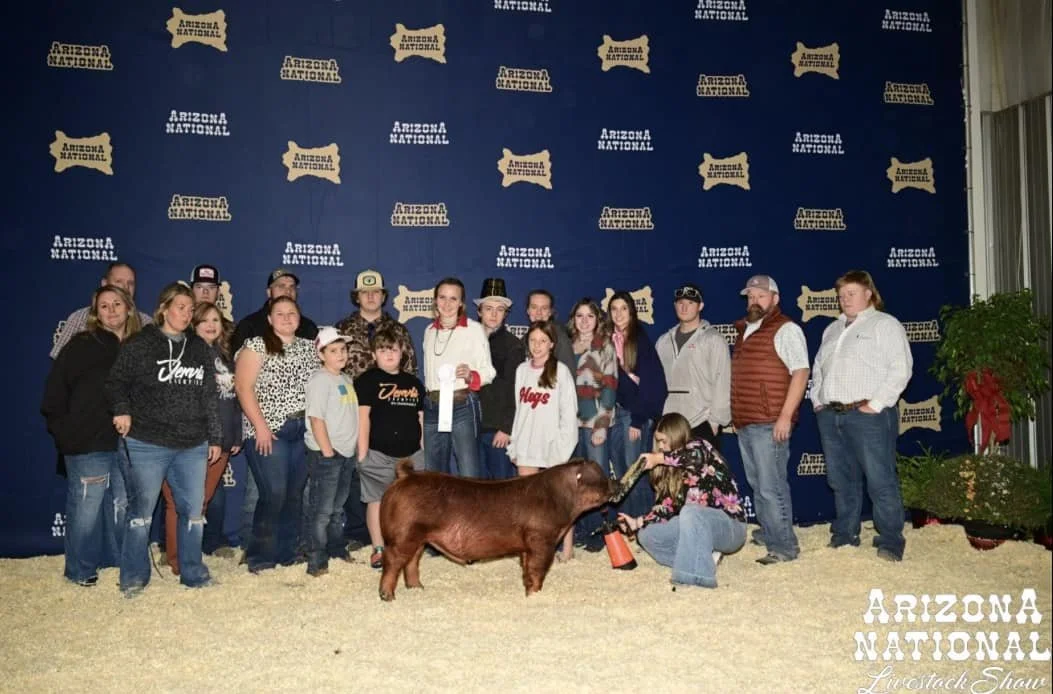 Group of people, including children, posing with a small brown cow at Arizona National Livestock Show in front of a backdrop with Arizona shape and event logo.